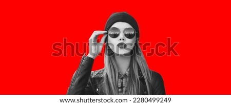 Fashionable portrait of stylish beautiful blonde young woman model blowing her lips with red lipstick sends sweet kiss in round sunglasses, black rock style leather jacket, hat