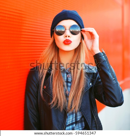 Fashionable portrait of stylish beautiful blonde young woman model blowing her lips with red lipstick sends sweet kiss in round sunglasses, black rock style leather jacket, hat on city street