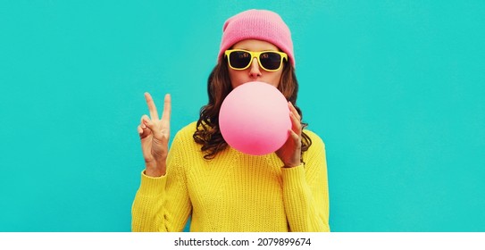 Fashionable portrait of stylish cool young woman inflating chewing gum wearing an yellow knitted sweater and pink hat on blue background