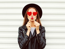 Fashionable Portrait Of Stylish Young Woman Covering Her Eyes With Red Heart Shaped Lollipop Blowing Her Lips Sending Sweet Kiss Wearing Black Round Hat, Leather Biker Rock Jacket On White Background