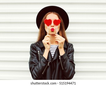Fashionable portrait of stylish young woman covering her eyes with red heart shaped lollipop blowing her lips sending sweet kiss wearing black round hat, leather biker rock jacket on white background