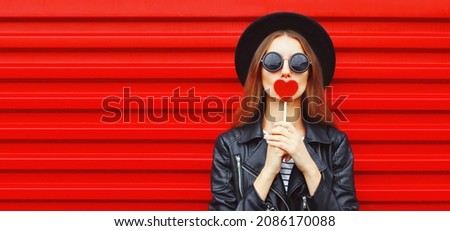 Fashionable portrait of stylish young woman with red heart shaped lollipop blowing her lips sending sweet air kiss wearing a black round hat, leather jacket on red background