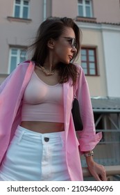 Fashionable Pretty Brunette Model Woman With Gold Chain Jewelry In Trendy Clothes Sits On The Street Near Building. Wearing A Pink Shirt, Tank Top, White Jeans And Black Purse