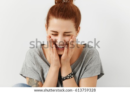 Fashionable pretty girl with hair knot and tattoo dressed in stylish grey t-shirt holding hands on cheeks and closing eyes, feeling embarassed, smiling shyly and joyfully sitting at blank studio wall