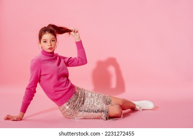 Fashionable redhead girl wearing trendy pink cashmere turtleneck sweater, sequin skirt, stylish white high top sneakers. Full-length studio portrait. Copy, empty space for text
