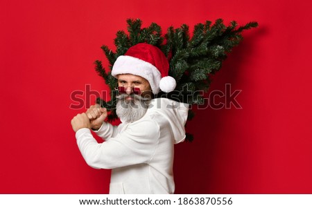 fashionable senior Santa with a hipster beard in a hat, white hoodie and stylish red glasses stands with a Christmas tree in his hands under the snow on a red background. Merry Christmas concept.