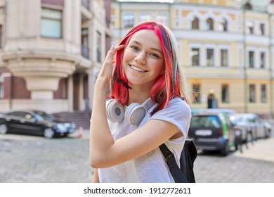 Fashionable smiling happy beautiful girl teenager 16, 17 years old with wireless headphones with bright dyed colored hairstyle on street of summer sunny city. Lifestyle, youth, fashion, beauty