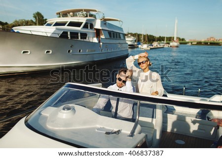 Fashionable stylish couple driving luxury motorboat yacht and posing in sunglasses with blue sky and yachts background