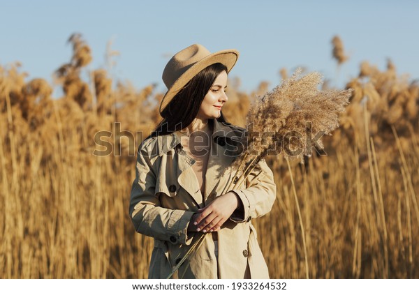 Fashionable and stylish girl in a beige trench coat\
and hat in the countryside on a background of dry reeds and blue\
sky in sunny day.