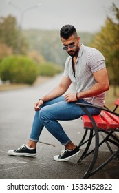 Fashionable Tall Arab Beard Man Wear On Shirt, Jeans And Sunglasses Sitting On Red Bench At Park.