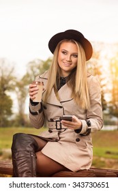 Fashionable Teenage Girl In Beige Trench Coat, Black Fedora Hat, And Boots Sitting On A Bench In Park Holding Smartphone And Cup Of Takeaway Coffee. Vertical, Retouched, Vibrant Colors.