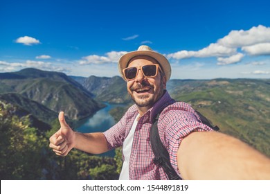 Fashionable tourist taking photo of himself while standing on hill above canyon and mountains and showing thumbs up. - Shutterstock ID 1544891819