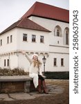 A fashionable woman in a beige winter outfit stands by a historic brick wall. She wears a cozy coat, beret, carrying a stylish brown handbag. Charming European architecture in the background