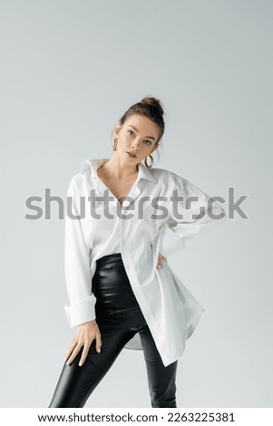 fashionable woman in black tight pants and white oversize shirt posing with hand on hip isolated on grey