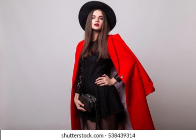 Fashionable Woman In Bright Red Coat And Black Hat Posing In Studio, Looking At Camera. Cold Season. Autumn Or Winter Look. 