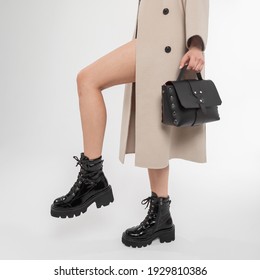 Fashionable Woman In A Classic Beige Coat With A Fashion Black Leather Bag In Stylish Shoes. Female Beautiful Legs With Trendy Boots In The Studio