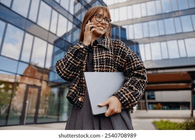 A fashionable woman engaged in a phone call while holding a laptop, set against a backdrop of modern architecture - Powered by Shutterstock