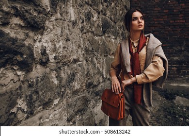 Fashionable Woman In Fashion Clothing. Beautiful Sexy Girl In Elegant Stylish Fall Clothes: Shirt, Scarf, Pants, Pullover, Leather Bag Posing Near Rock Wall Outdoors In Street In Autumn Or Spring