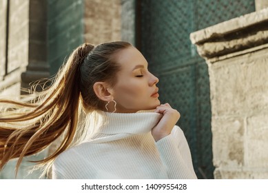 Fashionable woman with long hair in ponytail hairstyle wearing trendy hoop earrings, white turtleneck sweater, posing in street of European city. Copy, empty space for text