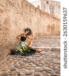 Fashionable woman is sitting on the stone street of beautiful historic town Calvi in island Corsica, France. Model wears elegant feminine outfit, long silk skirt and hat, looking into camera.