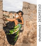 Fashionable woman is sitting on the stone wall of beautiful historic town Calvi in island Corsica, France. Model wears elegant feminine outfit, long silk skirt and hat during travels.