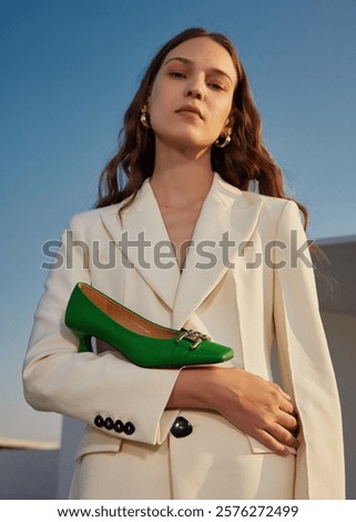 Fashionable woman in stylish white suit holding bold green shoe under blue sky.