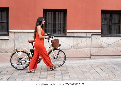 A fashionable woman in a trendy red jumpsuit strolls gracefully with her eyecatching bicycle in front of a vibrant wall, embodying urban fashion and a lively contemporary lifestyle - Powered by Shutterstock