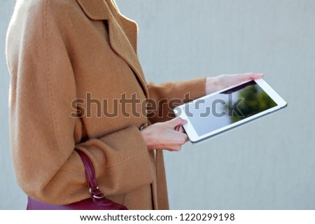 Fashionable woman wearing beige coat with burgundy bag and tablet gadget in hands. Freelance. Street style.