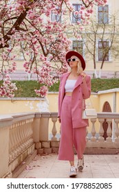 Fashionable woman wearing elegant pink suit, white crop top, sunglasses, burgundy hat, zebra print sneakers, holding white leather bag, walking in street near blooming magnolia tree