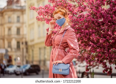 Fashionable Woman Wearing Spring Outfit With Blue Protective Face Mask, Pink Trench Coat, Small Bag. Model Posing Street Of European City. Trends During Quarantine Of Coronavirus Outbreak. Copy Space