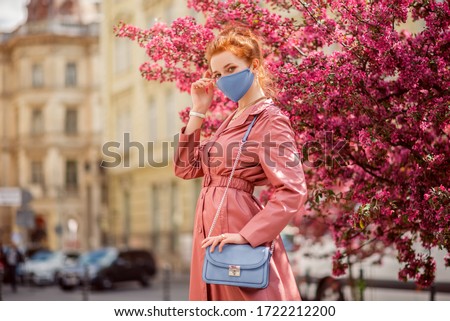 Fashionable woman wearing spring outfit with blue protective face mask, pink trench coat, small bag. Model posing street of European city. Trends during quarantine of coronavirus outbreak. Copy space