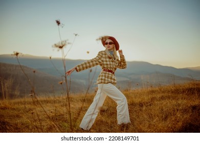 Fashionable Woman Wearing Stylish Autumn Outfit With Orange Hat, Sunglasses, White Wide Leg Jeans, Checkered Blazer, Belt, Walking In Mountain Nature During The Sunset. Copy, Empty Space For Text