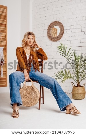 Fashionable woman wearing stylish boho outfit with suede jacket, wide leg jeans, sandals, holding trendy round wicker straw bag, handbag, posing in cozy room with green plants. 