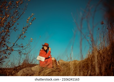 Fashionable Woman Wearing Stylish Orange Trench Coat, Hat, Brown Leather High Boots, With White Baguette Bag, Sitting, Posing In Nature. Outdoor Full-length Autumn Fashion Portrait. Copy, Empty Space
