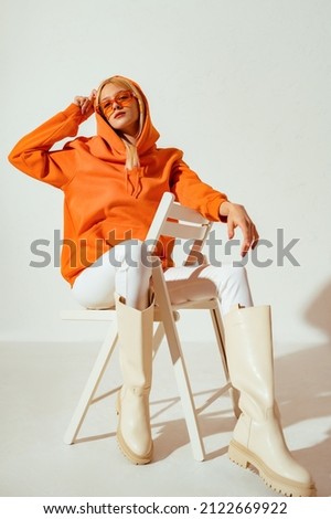 Fashionable woman wearing stylish orange hoodie, sunglasses, white skinny jeans, high boots sitting, posing on chair. Full-length studio portrait