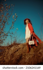 Fashionable Woman Wearing Stylish White Mini Dress, Orange Trench Coat, Hat, Brown Leather High Boots, Holding Handbag, Posing In Nature. Outdoor Full-length Autumn Fashion Portrait
