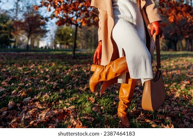 Fashionable woman wearing stylish white knitted dress, knee high boots with handbag in park. Fall shoes and accessories. Close up of boots
