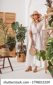 Fashionable woman wearing summer white crochet jumpsuit, straw hat, sandals, holding wicker bag, posing at home, in stylish boho interior with green tropical plants. Full-length indoor portrait
