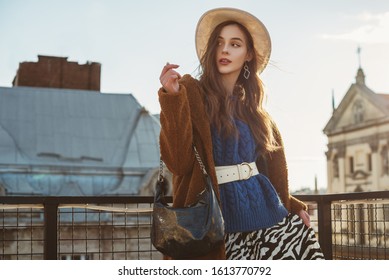 Fashionable  woman wearing trendy brown faux fur coat, blue knitted sweater, wide leather belt, stylish white hat, holding hobo handbag, posing at sunset, in European city. Copy, empty space for text