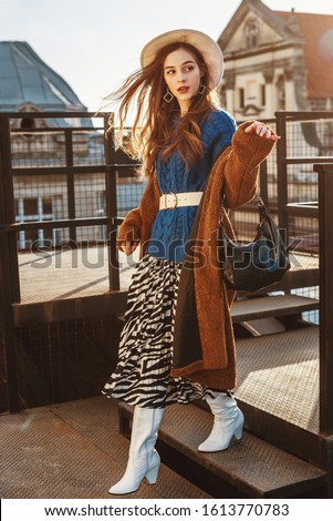 Fashionable  woman wearing trendy brown faux fur coat, blue knitted sweater, wide belt, stylish  hat, pleated zebra print skirt, white high boots, holding hobo bag, posing at sunset, in European city