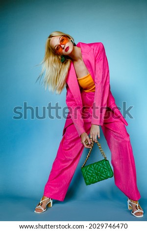 Fashionable woman wearing trendy fuchsia color suit. orange sunglasses, holding stylish green quilted faux leather bag with chunky chain, posing on blue background. Full-length studio fashion portrait