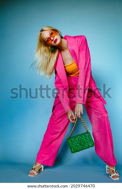 Fashionable woman wearing trendy fuchsia color\
suit. orange sunglasses, holding stylish green quilted faux leather\
bag with chunky chain, posing on blue background. Full-length\
studio fashion\
portrait