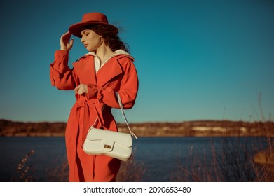 Fashionable Woman Wearing Trendy Orange Trench Coat, Hat, Holding White Textured Leather Handbag, Posing In Nature. Outdoor Autumn Fashion Portrait. Copy, Empty Space For Text