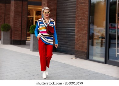 Fashionable Woman Wearing Trendy Outfit, Red Trousers, Blue Jacket, Striped Vest, White Shoes And Yellow Sunglasses. Model With Short Hair Walking The Street