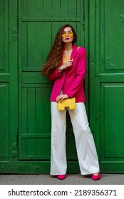 Fashionable Woman Wearing Trendy Outfit With Yellow Sunglasses, Bag, Pink Blazer, Wide Leg White Trousers, Posing Near Green Door. Full-length Outdoor Portrait