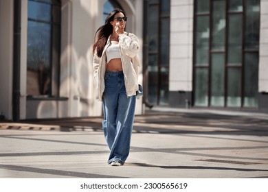 Fashionable woman wearing trendy white bomber jacket, CROP TOP, wide jeans walks on urban city street. Trends clothing of spring and summer.