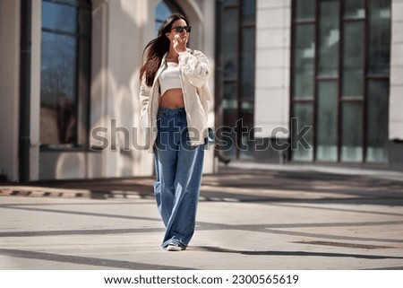 Fashionable woman wearing trendy white bomber jacket, CROP TOP, wide jeans walks on urban city street. Trends clothing of spring and summer. 商業照片 © 