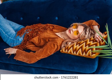 Fashionable Woman Wearing Trendy Yellow Sunglasses, Boho Style Faux Suede Jacket With Fringe, Laying On Blue Sofa