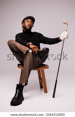 Fashionable young african american man in flat cap and glove holding golf club and sitting on chair on grey background