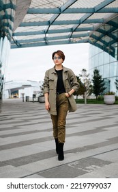 Fashionable Young Asian Woman With Short Hairstyle In Green Suit Walking In Outdoor Business District. Stylish Young Woman Wearing Glasses And Looking Away. Vertical View.